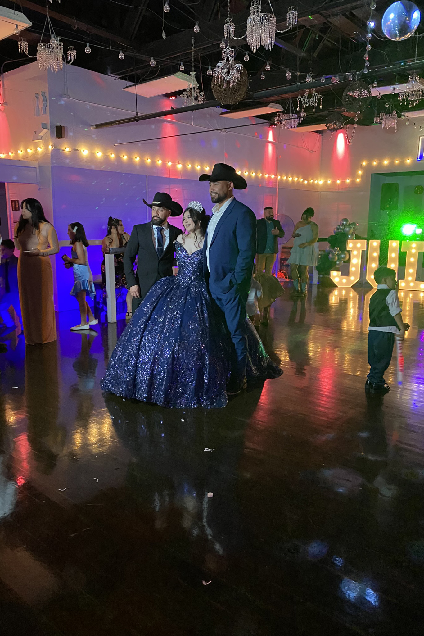 Two proud fathers posing with the quince girl of honor on dance floor in a throne chair.